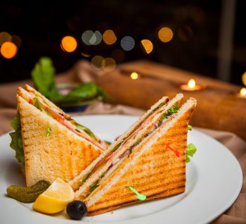 Side view club sandwich with salted cucumbers and lemon and olives in round white plate on wooden background